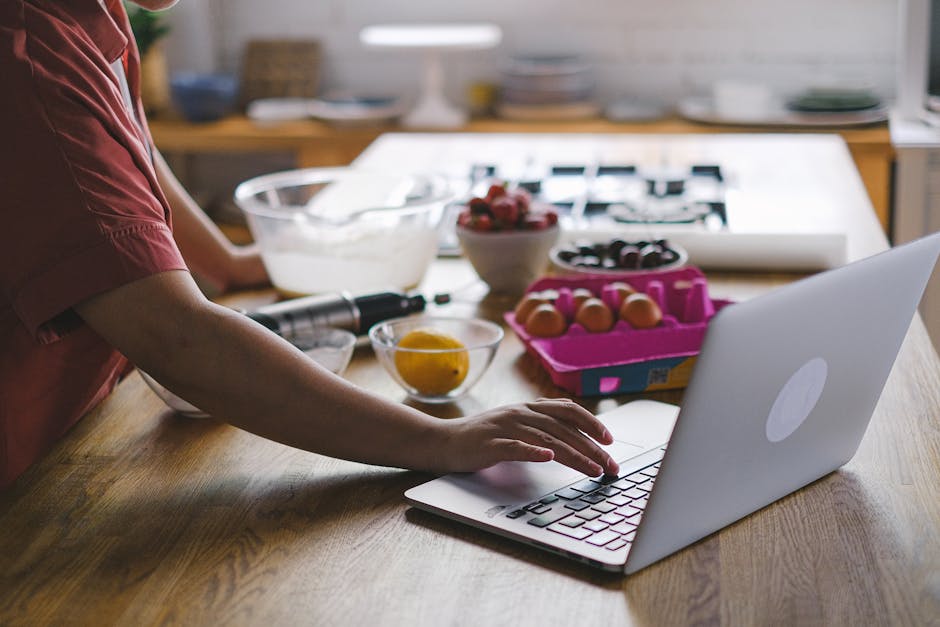 Person using a laptop in the kitchen while preparing baking ingredients. Modern multitasking lifestyle.