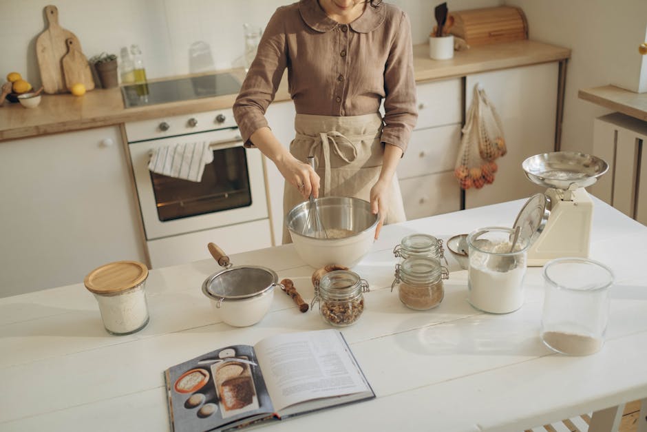 A person preparing dough in a cozy kitchen with various baking ingredients and a cookbook.