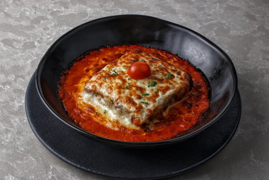 Tasty baked lasagna with cherry tomato and cheese, served in a black bowl with rich tomato sauce.