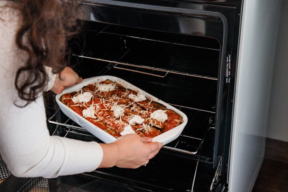 Woman placing a tray of lasagna into an oven with fresh cheese topping.