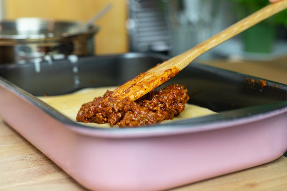 Close-up of layering meat sauce on lasagna in a pan in a kitchen setting.