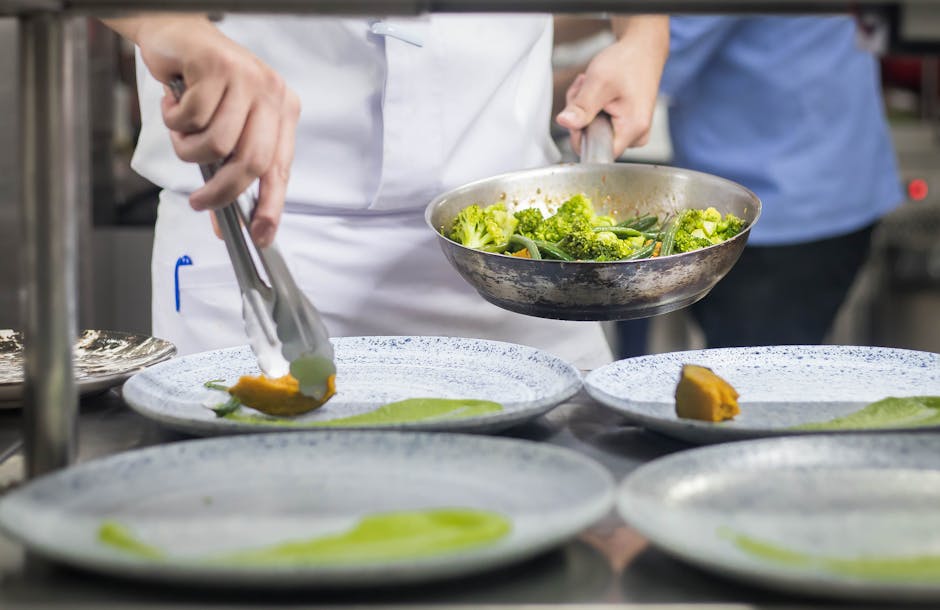 Chef plating a dish with broccoli and vegetables in a professional kitchen setting.