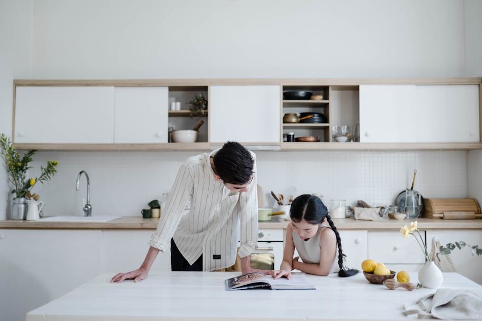 A father and daughter explore a cookbook together in a sleek, minimalist kitchen setting.